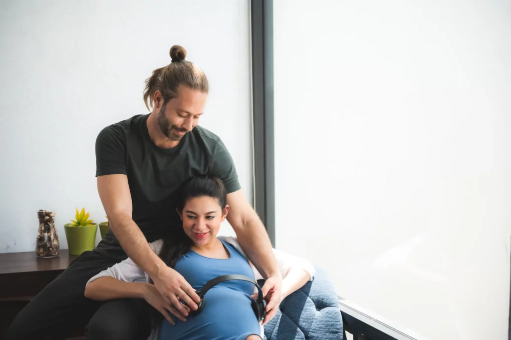 Smiling couple bonding during pregnancy, with headphones on pregnant belly to play music for unborn baby in bright modern home.