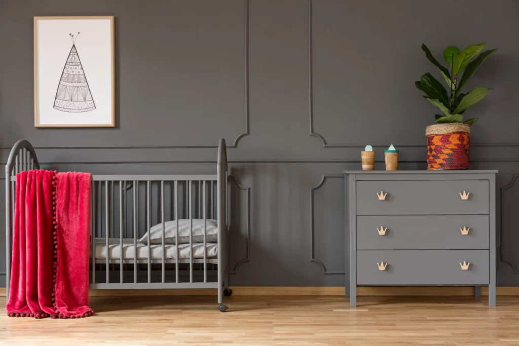 Modern gray baby nursery with gray crib, red plush blanket, and matching gray dresser with crown-shaped drawer pulls, accented by a vibrant potted plant and playful decor on a dark paneled wall with light wood flooring.
