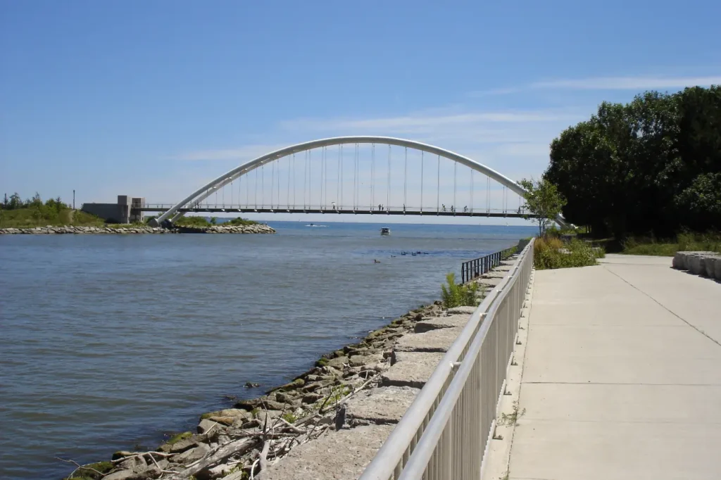 Toronto waterfront trail at Humber River pedestrian arch bridge with lakeside walkway, railing, calm water, and blue sky in Toronto Canada