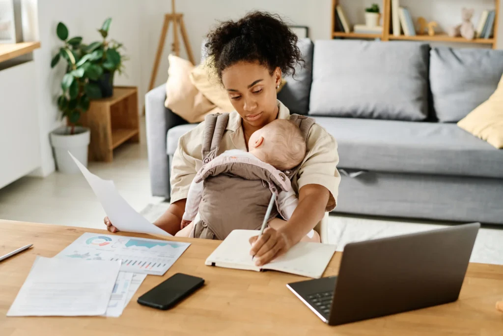 Young mother working from home with baby in carrier, multitasking with laptop, financial documents, and notepad, representing trying to calculate her EI maternity leave benefit in Canada