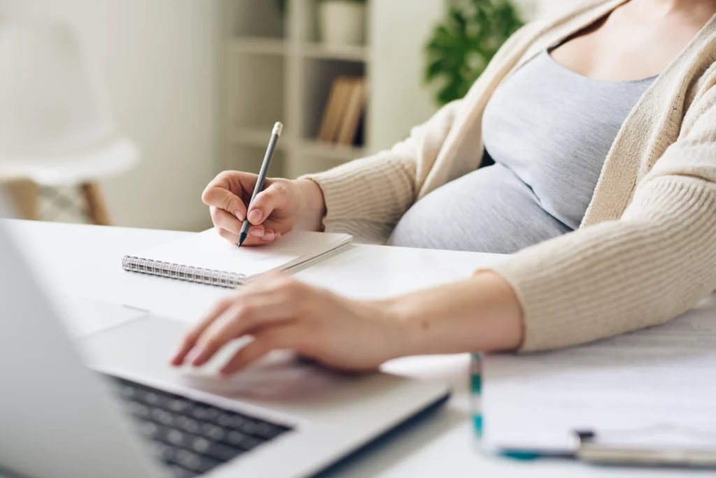Pregnant woman budgeting for maternity leave and EI benefits in 2026 using planner and laptop