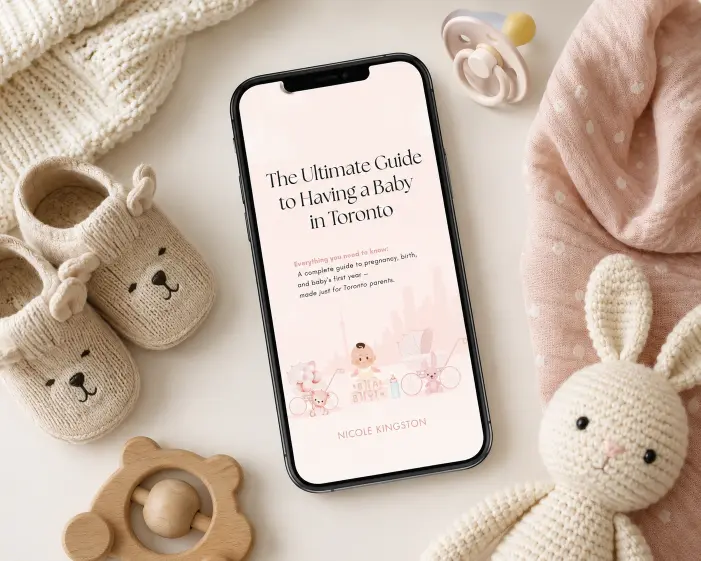 Smartphone displaying ‘The Ultimate Guide to Having a Baby in Toronto’ surrounded by cozy baby essentials including knitted booties, a pacifier, a plush bunny, and a wooden teething toy on a soft neutral background.