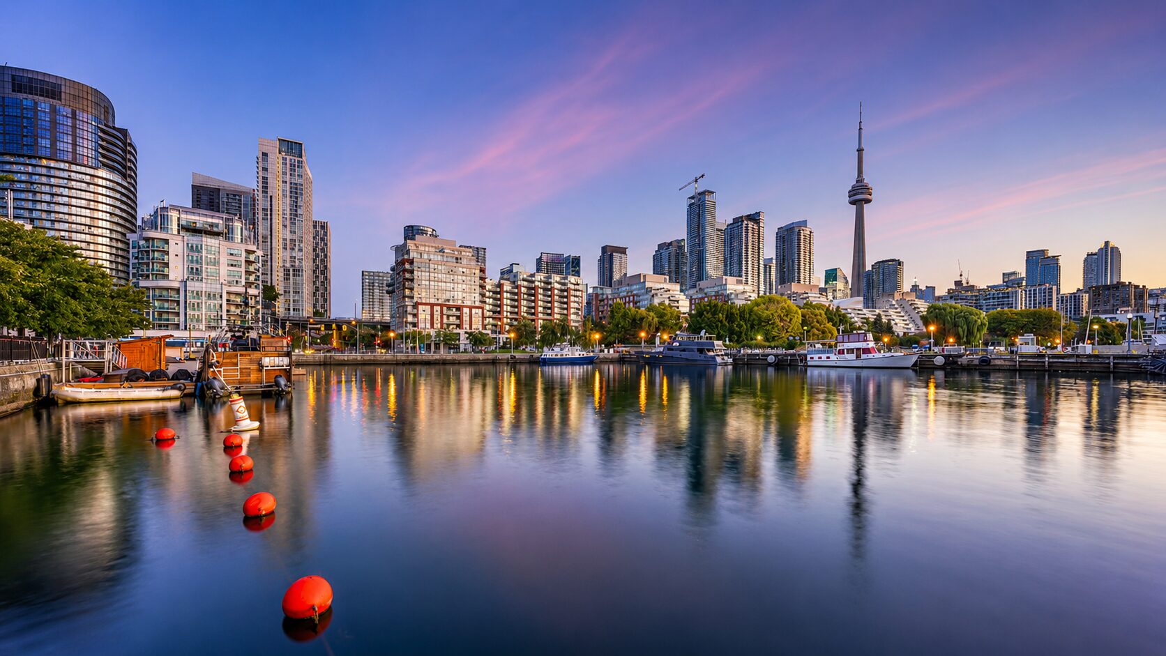 Toronto skyline and CN Tower viewed from Harbourfront at sunset with boats and waterfront reflections.